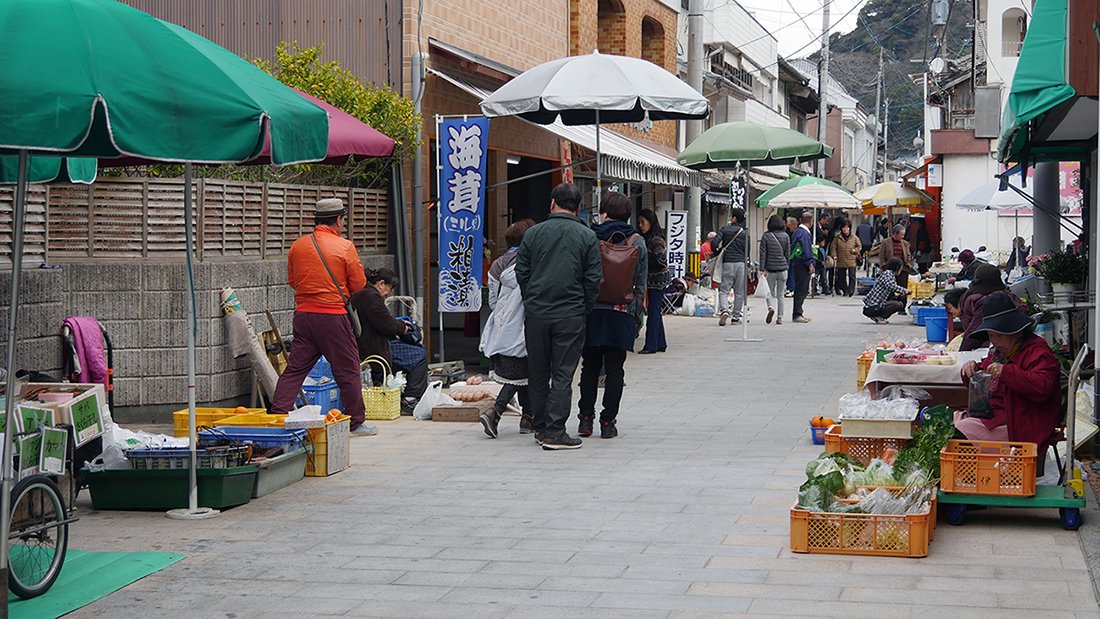 呼子朝市の朝市通り。パラソルを立てた露店が道の両側に並び、買い物客が行き交っている。